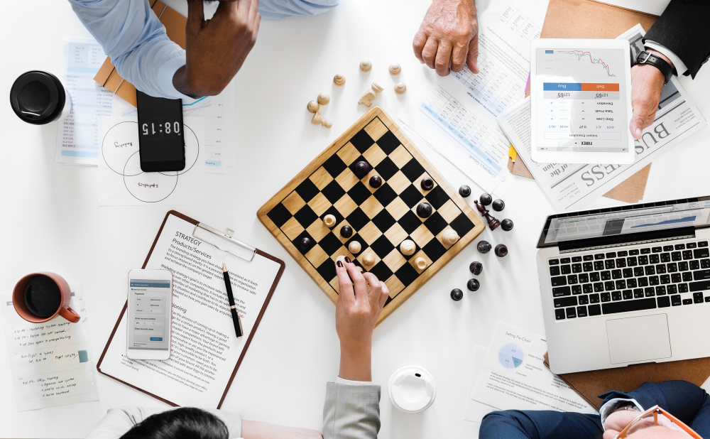 Business people scattered around a table playing a game of chess, strategic reports accompany