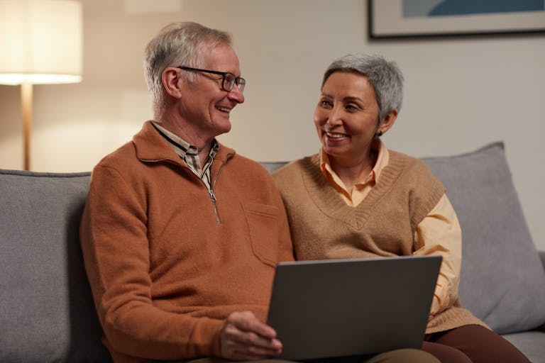 Senior couple smiling and using a laptop together in a cozy living room.