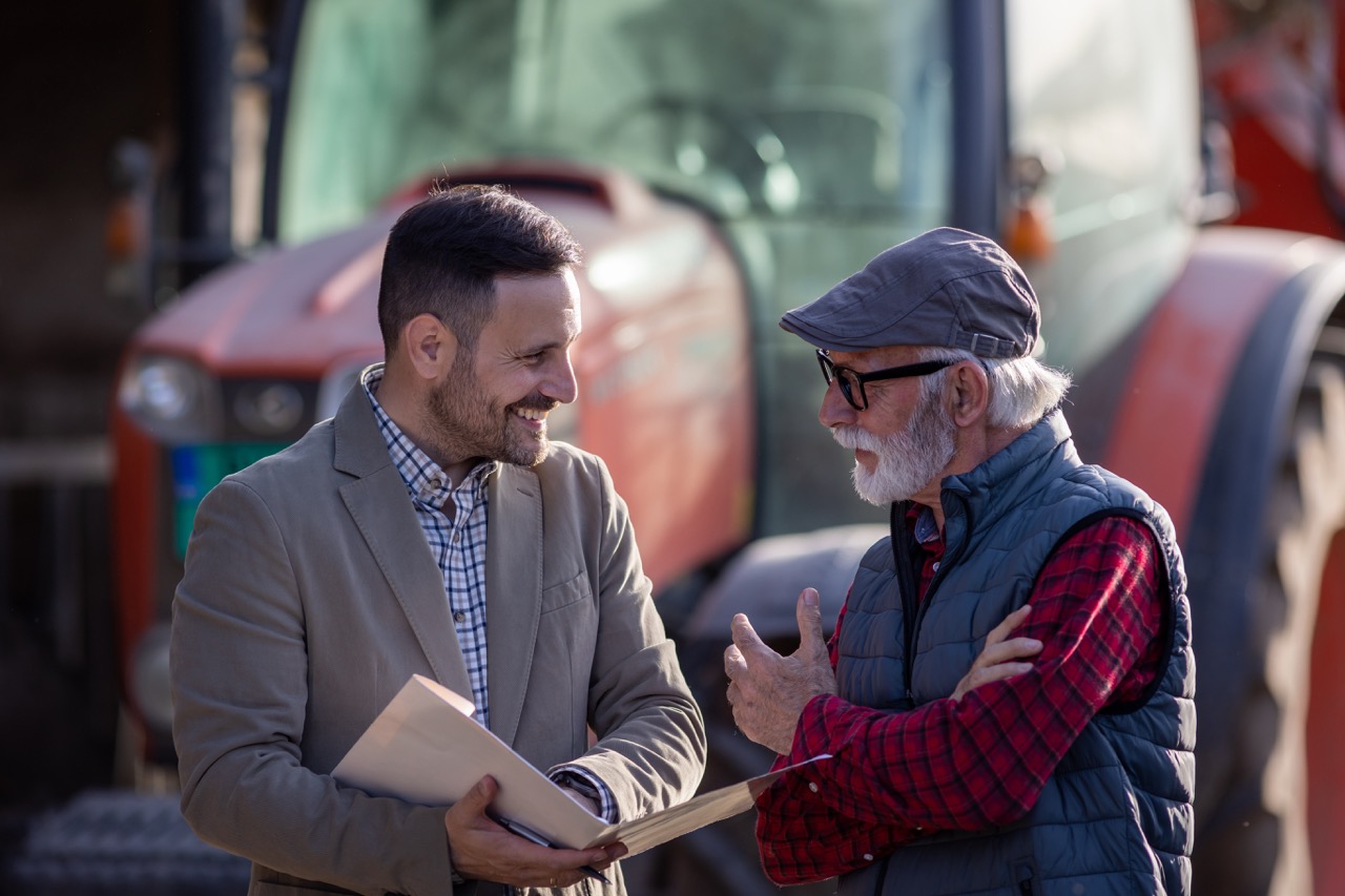 Smiling consultant and senior farmer talking about loan contract with bank in front of tractor on ranch